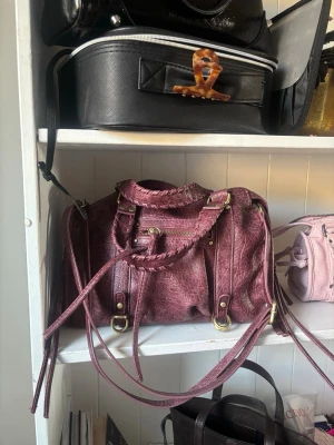 Vinröd handväska med flätade detaljer - Knappt använd, super bra skick. Inga fläckar eller skavanker. Snygg vinröd handväska med flätade handtag och långa dekorativa remmar. Väskan har guldfärgade metalldetaljer, dragkedja framtill och en mjuk, lite skrynklig läderlook. Perfekt för dig som gillar statement-accessoarer.