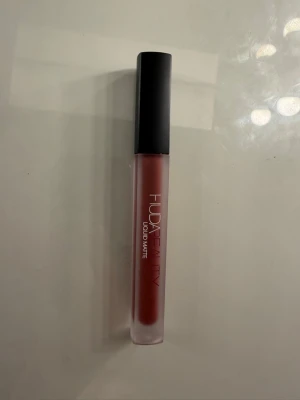 Huda Beauty Liquid Matte läppstift färg first class - Flytande läppstift från Huda Beauty i en snygg matt finish. Färgen är en varm, dämpad rosa-nude som passar perfekt till en trendig look. Kommer i en smal tub med svart lock.