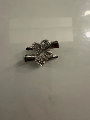 Silverfärgade hårspännen med hjärtan - Två hårspännen i silverfärg med glittrande strasstenar formade som hjärtan. Perfekta för att ge frisyren en söt och glammig touch. Metallmaterial med dekorativa stenar som fångar ljuset snyggt.
