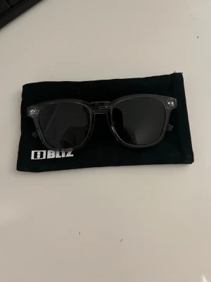 Solglasögon - Snygga gråa solglasögon från Bliz med klassisk fyrkantig form och transparenta bågar. Kommer med en svart tygpåse med Bliz-logga. Perfekta för att ge din stil en cool touch i solen.