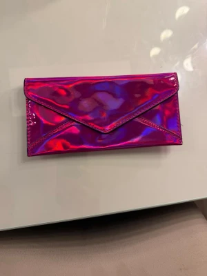 Holografisk rosa plånbok i konstläder - Cool plånbok i holografiskt rosa konstläder med kuvertformad framsida och dragkedjeficka på baksidan. Plånboken har en metallring för nycklar eller dekoration. Perfekt för dig som gillar färg och vill sticka ut.
