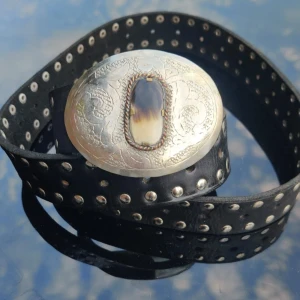 Svart bälte med stor silverspänne - Coolt svart bälte i skinn med silverfärgade nitar längs hela remmen och ett stort, ovalt silverspänne med ingraverat mönster och en dekorativ sten i mitten. Perfekt accessoar för att lyfta din outfit.