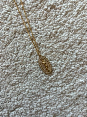 Guldigt halsband med oval berlock - Snyggt halsband i guldfärgad metall med en tunn kedja och en oval berlock. Berlocken har en upphöjd figur i mitten och är dekorerad med små gnistrande stenar runt kanten. Perfekt accessoar för att lyfta din outfit.
