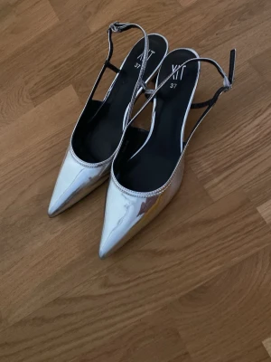 Silvriga pumps med rem från XIT - Säljer ett par glansiga silvriga pumps från XIT i storlek 37. Skorna har spetsig tå, öppen häl och justerbar rem runt ankeln. Perfekta för dig som vill sticka ut med en futuristisk look.oanvända 