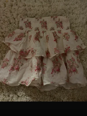 Blommig volangkjol från Kappahl - Supersöt kort kjol från Kappahl med blommigt mönster i rosa och grönt på vit botten. Kjolen har bred smock i midjan och flera volanger som ger en romantisk och luftig känsla. Perfekt för vår och sommar!