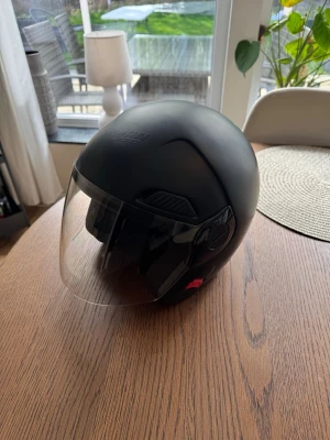 Svart MC-hjälm med visir - Snygg svart MC-hjälm från Nolan med stort genomskinligt visir och vadderade öronkuddar. Hjälmen har en rundad form och är tillverkad i tåligt material, perfekt för dig som kör moped eller motorcykel och vill ha både stil och skydd.