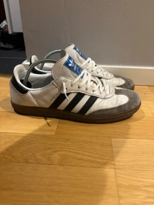 Adidas Samba vit - Skorna har en liten röd prick på framsidan som är i turs vilket går att få bort med en skrubb. Klassiska Adidas Samba sneakers i vitt med svarta ränder på sidorna och blå logga på plösen. Ovandelen är i mocka och textil, yttersulan är brun. Tidlös retrostil med låg profil och snörning framtill.