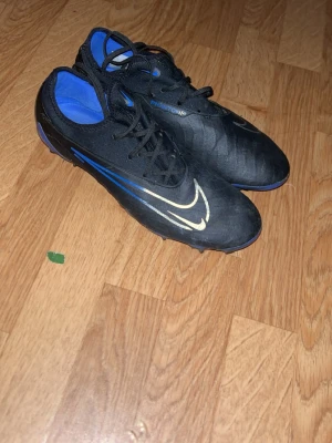 Nike Phantom GX PRO FG SHADOW - Säljer ett par Nike Phantom GX PRO FG SHADOW  fotbollsskor i svart med blå detaljer och vit Nike-logga. Skorna har snörning, låg profil och är tillverkade i syntetmaterial med textilfoder. Perfekta för dig som vill ha kontroll och stil på planen.