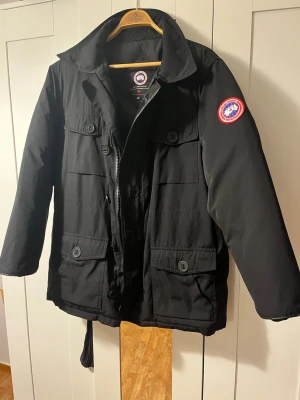 Svart parkas från Canada Goose - Nästan som ny! Snygg svart parkas från Canada Goose med klassisk logga på ärmen. Jackan har flera stora fickor med lock och knappar, dragkedja framtill och hög krage. Storlek L men passar M. Och den passar dam och herr.