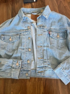 Ljusblå Levi's jeansjacka - Klassisk ljusblå jeansjacka från Levi's med raka linjer, två bröstfickor med lock och knapp, samt knappstängning framtill. Jackan har en avslappnad passform och ikonisk röd Levi's-tag på bröstfickan. Perfekt för en cool och tidlös look, endast använd 1 gång.
