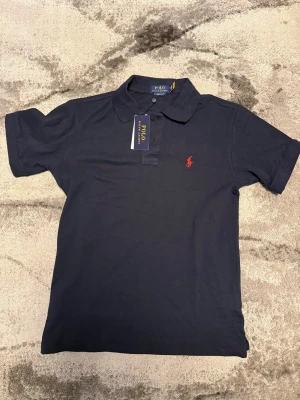 Mörkblå pikétröja från Polo Ralph Lauren - Klassisk mörkblå pikétröja från Polo Ralph Lauren med röd broderad logga på bröstet. Tröjan har krage, korta ärmar och knäppning framtill. Perfekt för en stilren och sportig look.