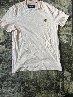 Ljusrosa t-shirt från Lyle & Scott - Säljer en ljusrosa t-shirt från Lyle & Scott med klassisk passform och korta ärmar. T-shirten har den ikoniska gula loggan broderad på bröstet och är tillverkad i mjuk bomull. Perfekt till jeans eller shorts för en clean look.