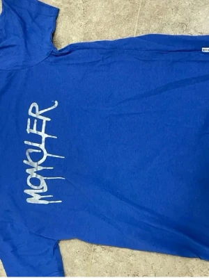 Blå Moncler t-shirt med tryck - Snygg blå t-shirt från Moncler med ett stort vitt tryck på bröstet i graffiti-stil. Klassisk passform och rund halsringning, perfekt för en avslappnad och trendig look.