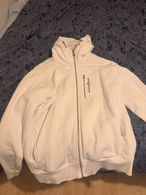 Vit hoodie från Sail Racing - Snygg vit hoodie från Sail Racing med dragkedja framtill och en extra ficka med zip på bröstet. Hoodien har justerbar huva med snörning och ribbade muddar. Perfekt för dig som gillar stilren och sportig look.