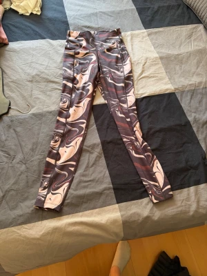 Mönstrade swirl-leggings i brunt och rosa - Säljer ett par snygga leggings med swirl-mönster i bruna, rosa och beige toner. De har hög midja och är i stretchigt material som sitter tight. Perfekta för dig som gillar unika och trendiga träningskläder eller vill ha något extra till vardagslooken.