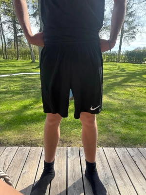 Svarta Nike shorts  - Säljer ett par svarta träningsshorts från Nike med klassisk vit logga på benet. Shortsen har elastisk midja och är tillverkade i lätt och bekvämt polyestermaterial, perfekta för sport och aktiva dagar. Små i storleken