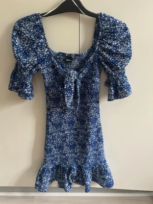 Blå blommig kortklänning med volang - Supersöt blå kortklänning från NA-KD med vita småblommor, puffärmar och volangkant nertill. Klänningen har en knytning framtill och är perfekt för sommarens alla tillfällen. Skön passform och lätt material.