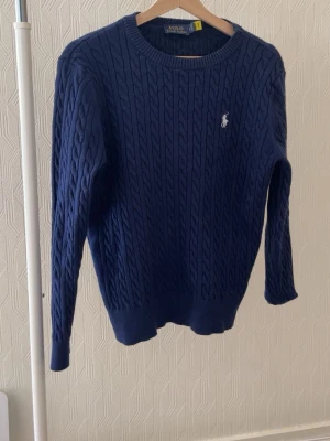 Mörkblå kabelstickad tröja Polo Ralph Lauren - Kabelstickad mörkblå tröja från Polo Ralph Lauren med rund halsringning och vit broderad logga på bröstet. Långärmad modell med ribbade muddar och nederkant. Perfekt för lager-på-lager och klassisk stil.