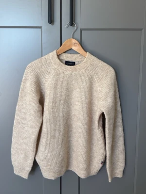 Beige stickad tröja från Lexington - Klassisk beige stickad tröja från Lexington med rund halsringning och ribbstickade muddar. Tröjan har en rak passform och är tillverkad i ett mjukt, ulligt material som känns både stilrent och bekvämt. Perfekt för lager-på-lager under kyliga dagar.