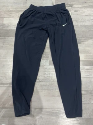 Svarta träningsbyxor från Nike - Snygga svarta träningsbyxor från Nike med vit logga på vänster lår. Byxorna har resår i midjan och dragkedjeförsedda fickor. Perfekta för gymmet eller löprundan, tillverkade i ett lätt och bekvämt material.