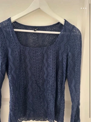 Blå spetsblus från ONLY - Supersnygg blå långärmad spetsblus från ONLY med fyrkantig ringning och genombrutet mönster. Perfekt att styla med jeans eller kjol för en trendig look. Blusen har en figurnära passform och fina spetsdetaljer över hela plagget.