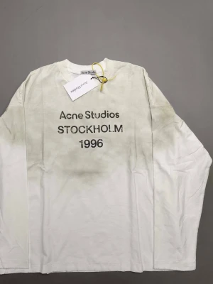 Vit långärmad topp Acne Studios - Vit långärmad topp från Acne Studios med tryck 'Acne Studios STOCKHOLM 1996' på bröstet. Rund halsringning och avslappnad passform. Perfekt för en cool och enkel stil.