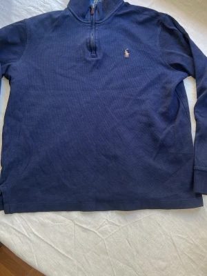 Marinblå halvzip från Polo Ralph Lauren - Klassisk marinblå långärmad tröja från Polo Ralph Lauren med halv dragkedja och broderad logga på bröstet. Tröjan har ribbade muddar och är tillverkad i ett mjukt bomullsmaterial. Perfekt för en stilren och avslappnad look.