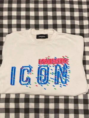Vit Dsquared2 ICON hoodie - Säljer en vit hoodie från Dsquared2 med färgglad 3D-tryckt 'ICON' och Dsquared2-logga på bröstet. Hoodien har rund hals, långa ärmar och ribbade muddar. Materialet är mjukt och bekvämt, perfekt för en trendig streetwear-look.
