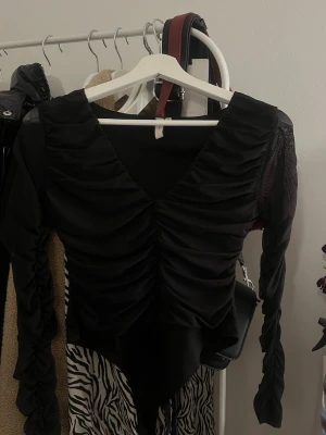 Svart mesh body med rynkade detaljer - Säljer en svart body med långa ärmar i mesh och snygga rynkade detaljer framtill. Djup V-ringning och figurnära passform. Materialet är en mix av mesh och stretchigt tyg som ger en cool look. Perfekt för dig som vill ha något trendigt och stilrent.