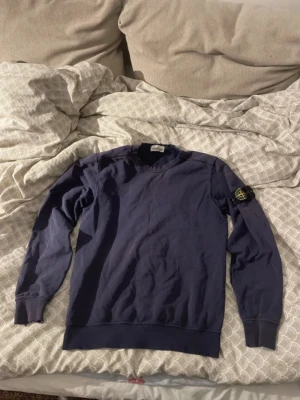 Mörkblå sweatshirt från Stone Island - Mörkblå sweatshirt från Stone Island Junior med klassisk rund halsringning och ikonisk logotyp-patch på vänster ärm. Tröjan har ribbade muddar vid ärmslut och nederkant. Perfekt för en avslappnad och stilren look.