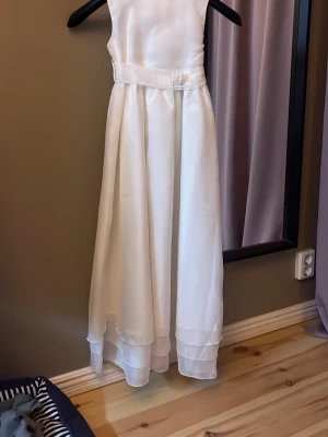 Vit långklänning  - Elegant vit långklänning i satinliknande material med markerad midja och avtagbart bälte med rosettdetalj. Klänningen är ärmlös och har en stilren, klassisk design med lätt utställd kjol och diskreta lager längst ner. Använd 1 gång på ett bröllop 