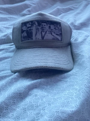 Grå keps med mesh och fototryck - Säljer en grå keps med mjuk textil och mesh på sidorna. På framsidan finns ett svartvitt fototryck med sporttema och en signatur. Kepsen har böjd skärm och justerbar baksida. Perfekt för dig som gillar unika accessoarer.