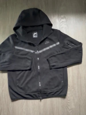 Svart Nike hoodie med dragkedja - Svart hoodie från Nike med hel dragkedja och huva. Har en diskret logga på bröstet och en grå diagonal detalj över framsidan. Praktisk ficka med dragkedja på ena ärmen. Perfekt för en sportig och avslappnad look.