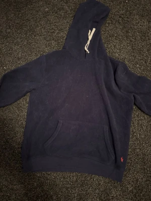 Mörkblå hoodie från Polo Ralph Lauren - Mörkblå Polo Ralph hoodie med huva . Klassisk känguruficka framtill och diskret röd logga broderad vid nederkanten. Perfekt för användning i vardagen och i vardagsrummet .