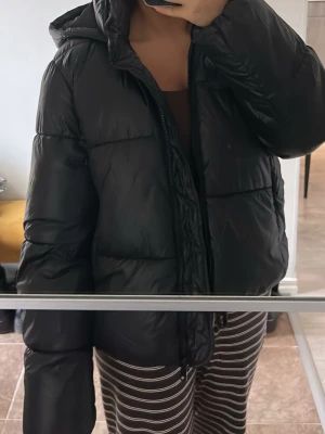 Svart pufferjacka  - Säljer en svart pufferjacka med huva och dragkedja framtill. Perfekt för vinter/höst och vår som är enkel att styla till olika outfits. Materialet är glansigt. Använd 2-5 gånger