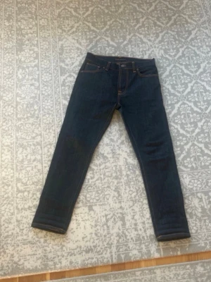 Mörkblå jeans från Nudie Jeans - Säljer ett par mörkblå jeans från Nudie Jeans i rak modell. Jeansen är tillverkade av ekologisk bomull och har klassiska orange sömmar samt fem fickor. Perfekta för dig som gillar stilrena och tidlösa jeans.