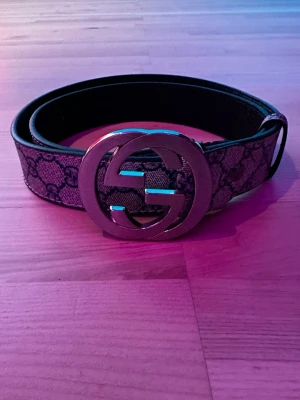 Gucci GG bälte med monogramspänne - Snyggt bälte från Gucci med det klassiska GG-monogrammet i mörka toner och ett stort, silverfärgat GG-spänne. Bältet har en exklusiv känsla och är tillverkat i skinn med canvasdetaljer. Perfekt accessoar för att lyfta vilken outfit som helst.