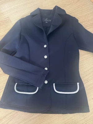 Marinblå kavaj från Exclusive - Snygg marinblå kavaj från Exclusive med fyra glittriga knappar framtill och vita detaljer vid fickorna. Figurnära passform och klassisk krage gör den perfekt till ridning eller tävling. Materialet känns stretchigt och bekvämt.