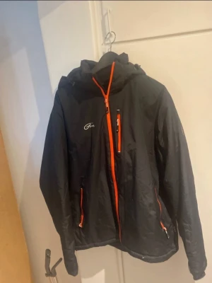 Svart vindjacka från Five med orange dragkedjor - Snygg svart vindjacka från Five med orange dragkedjor och detaljer. Jackan har huva, tre fickor med dragkedja och logga på bröstet. Tillverkad i lätt och vindtätt material, perfekt för blåsiga dagar.