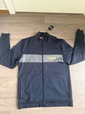 Mörkblå zip hoodie från BOSS - Snygg mörkblå hoodie från BOSS med dragkedja framtill och en grå rand över bröstet där BOSS-loggan syns i gult. Hoodien har två fickor fram och ribbade muddar vid ärmslut och nederkant. Tillverkad i mjukt material som känns bekvämt mot huden.