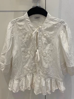 Vit blus med spetsdetaljer och volang - Söt vit blus från Pimkie med spetsdetaljer framtill, knytband och volangkant nedtill. Blusen har trekvartsärm och puffiga axlar för en romantisk look. Tillverkad i ett lätt bomullstyg som känns luftigt och bekvämt. Köpt i Fransk affär i Frankrike 