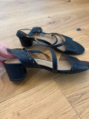 Svarta sandaler med klack från Caprice - Snygga svarta sandaler från Caprice med låg blockklack och justerbara remmar. Sandalerna har dekorativa håldetaljer och öppen tå. Tillverkade i skinn med mjuk innersula för extra komfort. Perfekta för dig som vill ha både stil och bekvämlighet.