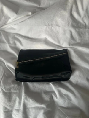 Svart necessär från Yves Saint Laurent - Snygg svart necessär från Yves Saint Laurent i glansigt material med guldfärgad dragkedja och YSL-logga på dragkedjans hänge. Perfekt för smink eller småprylar. Enkel, stilren design med rektangulär form. Längd: ca 25cm höjd: ca 15cm bredd: ca 5,5cm
