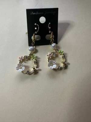 Blommiga örhängen med pärlor blommor och bin - Säljer ett par unika örhängen med blommor och bin med guldfärgade ringar, vita och rosa blommor samt gröna detaljer. Örhängena har även en vit pärla som hänger ovanför blommorna. Perfekt för dig som gillar färgglada accessoarer.