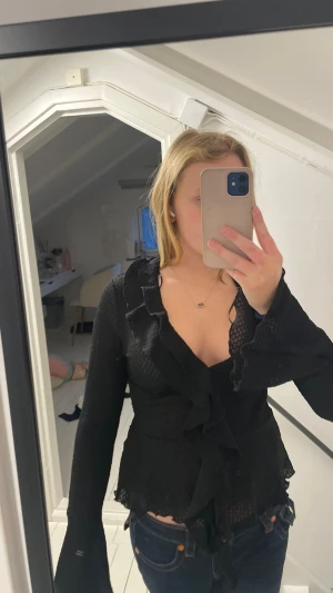 Svart volangblus från Gina Tricot - Säljer en svart blus från Gina Tricot med volangdetaljer längs fram och vid ärmslut. Blusen har långärmad design och är tillverkad i ett lätt, transparent material som ger en cool och trendig look. Perfekt att styla med jeans eller kjol.