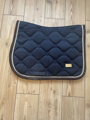 Marinblått schavrak från equstrian Stockholm  - Snyggt marinblått schabrak från Equestrian Stockholm  med quiltat mönster och guldfärgad kant. Schabraket har en liten metallbricka med logga och är kantat med brunt tyg och tvinnad snodd i guld och vitt. Perfekt för dig som vill ha stil på ridturen.