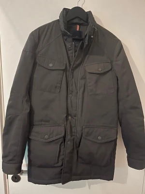 Mörkgrå parkajacka från Riley - Snygg mörkgrå parkajacka från Riley med fyra stora fickor framtill och hög krage. Jackan har långa ärmar, knappar och dragkedja framtill samt en klassisk, rak passform. Perfekt för kyligare dagar och enkel att matcha med olika outfits.