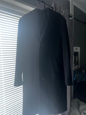 Svart klassisk ullkappa - Stilren svart kappa med tre knappar framtill och klassisk krage. Kappan har raka ärmar och två diskreta fickor på sidorna. Perfekt för dig som vill ha en tidlös och enkel look. Materialet ser ut att vara ullblandning. Helt NY