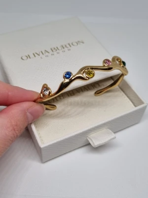 Guldpläterad armband - Snyggt guldfärgat armband med vågig form och infattade färgglada stenar i blått, gult, rosa, grönt och vitt. Armbandet är öppet och har en modern, lekfull design som sticker ut. Lådan ingår ej.