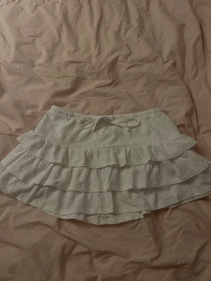 Vit volangkjol med spetsdetaljer - Supersöt vit kortkjol med flera volanger och broderade spetsdetaljer. Kjolen har en liten rosett framtill och elastisk midja för skön passform. Perfekt för dig som gillar romantisk och feminin stil.